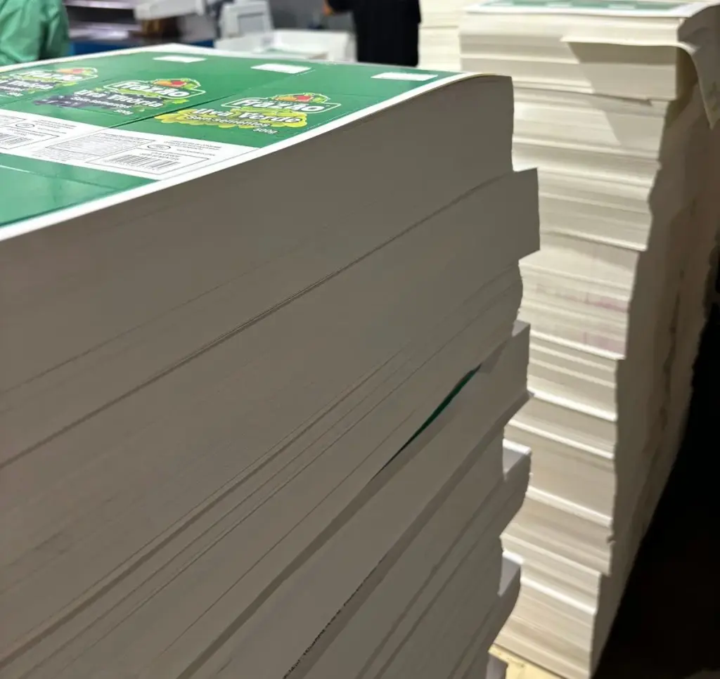 Pilhas de folhas impressas para cintas em cartão triplex C1S 190 g, com corte especial e colagem, e um operador da iGráfica ao fundo