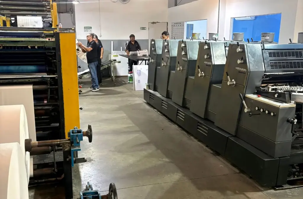 Corredor da fábrica da iGráfica com a impressora offset Heidelberg de cinco unidades à direita e a máquina de bobina à esquerda, com a equipe trabalhando ao fundo