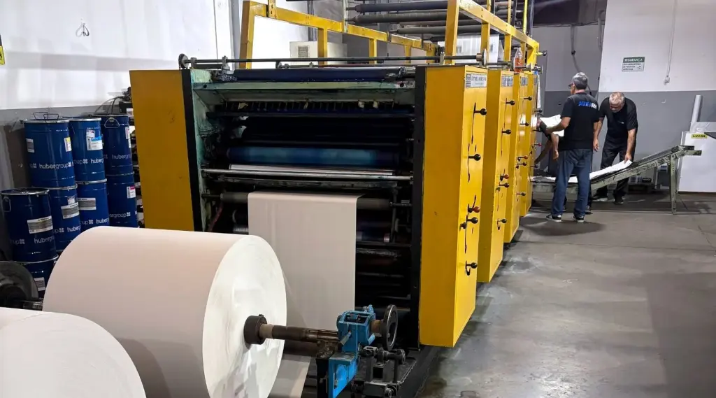 Rotativa Harris Cottrell V-15 da iGráfica, com bobinas de papel em primeiro plano e dois operadores ao fundo