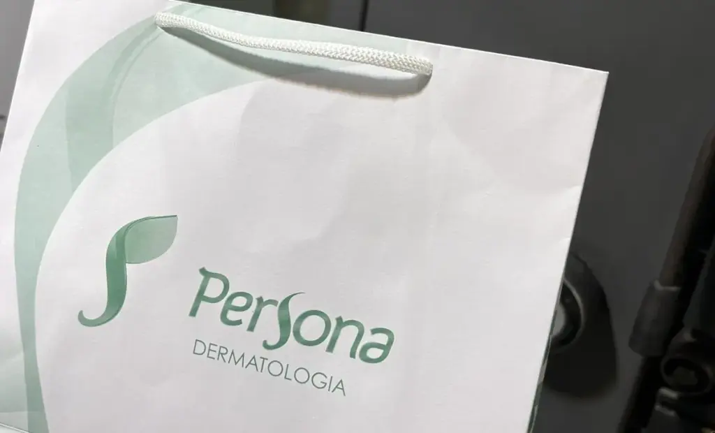 Sacola de papel branca com alça de nylon e a arte em tons de verde da Persona Dermatologia, impressa pela iGráfica