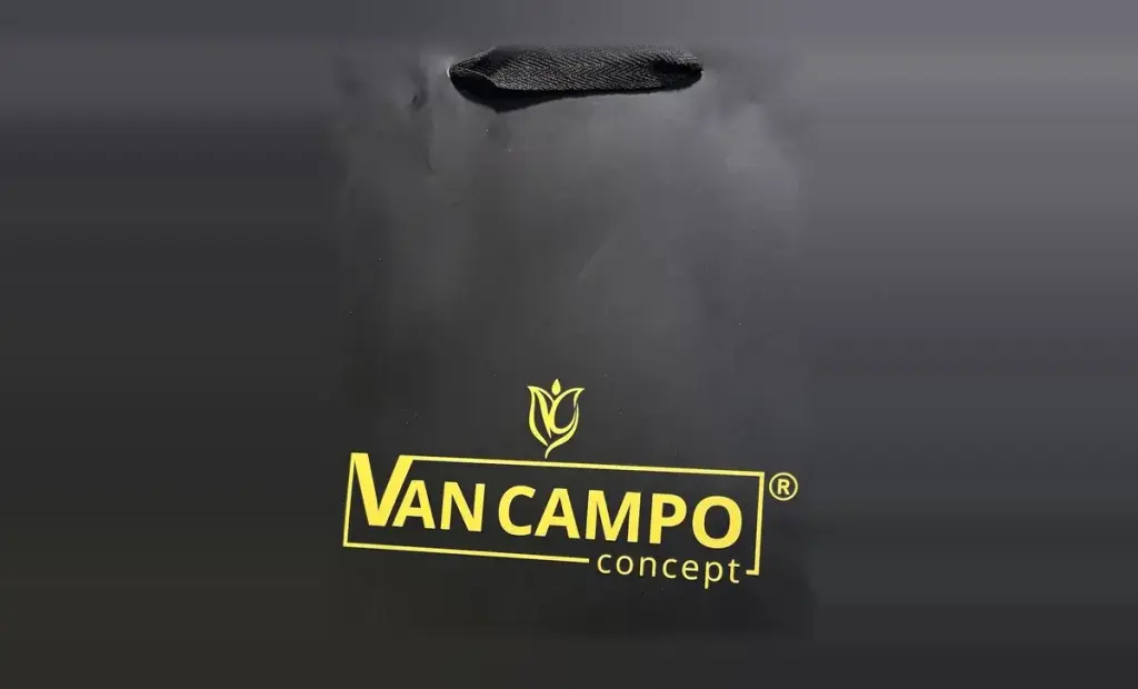 Sacola de papel da Van Campo Concept, impressa em cores, com fundo preto, logomarca em amarelo e alça de fita preta